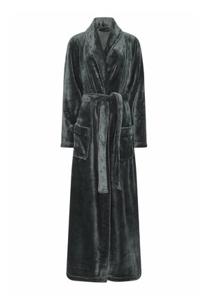 Femme portant un long peignoir en peluche gris foncé avec une ceinture nouée à la taille et les mains dans les poches avant, debout devant un fond uni.