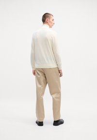 Pull crème à col montant, fabriqué en matériau doux, associé à un pantalon beige clair et des chaussures noires. Design simple et décontracté.