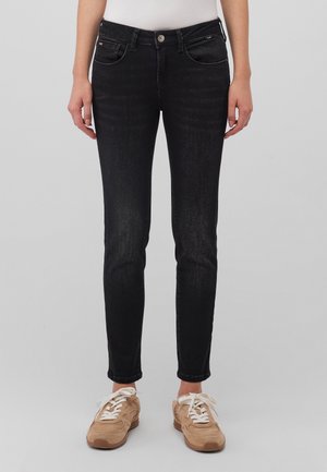 Henkilö, jolla on päällään kapeat mustat farkut ja beigejä lenkkareita, seisoo yksinkertaisen harmaan taustan edessä.