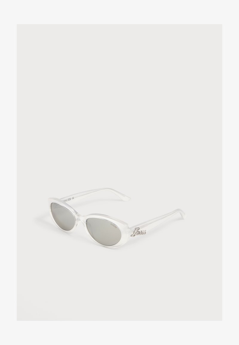 Lunettes de soleil œil de chat avec montures transparentes claires et verres teintés gris, avec un logo métallique "Guess" sur la branche.