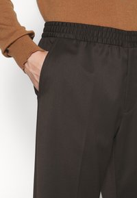 Main dans la poche d'un pantalon marron foncé à taille élastique, porté avec un haut marron à manches longues en maille côtelée avec des poignets festonnés.