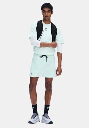 Coureur masculin portant un short de sport bleu clair et un t-shirt, un gilet d'hydratation noir, des chaussettes noires et des chaussures de course blanches, debout avec un bras croisé sur la poitrine.