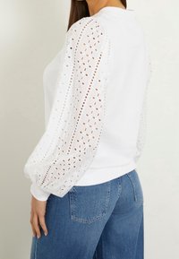 Sudadera blanca con detalle de ojales bordados en las mangas abullonadas, con escote y puños acanalados, combinada con vaqueros de talle alto.