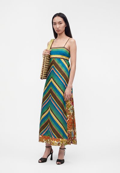 Γυναίκα που φοράει ένα αμάνικο, ριγέ maxi φόρεμα με λουλουδάτες λεπτομέρειες, κρατώντας μια πλεκτή τσάντα tote και φοράει μαύρα ανοιχτά πέδιλα.