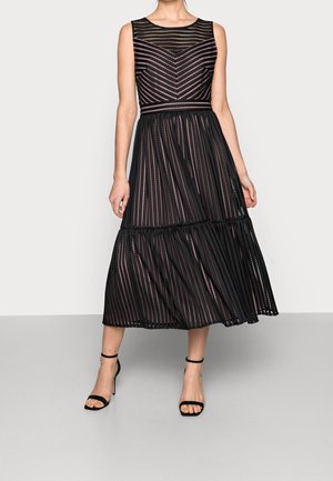 Sort ærmeløs midi-kjole med chevron- og stribet mesh-overlay, figursyet talje og flæset nederdel i lag, kombineret med sorte ankelrem-hæle.