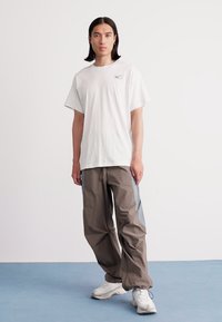 Young person with long dark hair stands wearing a white Nike t-shirt, gray and brown loose pants, and white sneakers on a blue floor.