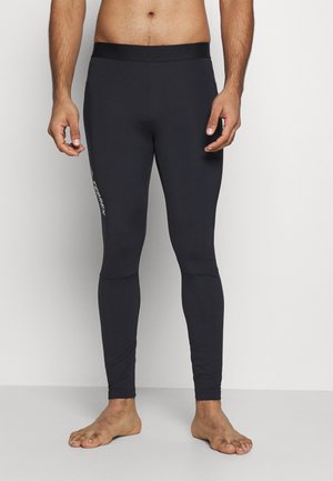 Mann trägt schwarze, eng anliegende Sportleggings mit „TERREX“-Logo am linken Oberschenkel, steht barfuß vor einem einfarbigen hellen Hintergrund.