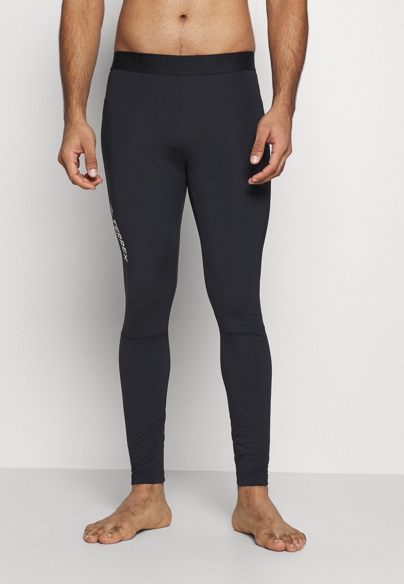 Homme portant un legging de sport noir ajusté avec le logo « TERREX » sur la cuisse gauche, debout pieds nus sur un fond clair uni.