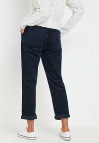 Mujer vestida con pantalones de pana azul marino arremangados en los puños, zapatillas blancas y un suéter de punto blanco, de pie con las manos en los bolsillos traseros.