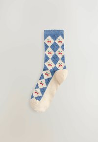 Blaue und cremefarbene Socke mit einem Rautenmuster, in dem kleine rote Kirschen in einem wiederkehrenden Design auf einem neutralen Hintergrund angeordnet sind.