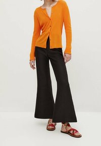 Oranger Rippstrick-Cardigan mit Knöpfen, schwarze ausgestellte Hose und mehrfarbige Riemchensandalen. Glatte Textur und lockere Passform.