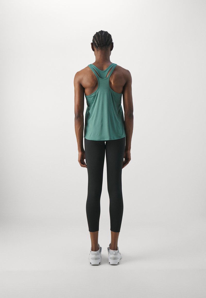 Canotta atletica verde con spalline incrociate sul retro, abbinata a leggings neri e scarpe sportive bianche, vista di spalle. Texture lisce.