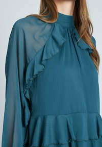 Blusa teal con colletto alto e dettagli arricciati sulle spalle e sul petto. Realizzata in tessuto leggero e testurizzato con una silhouette fluida.