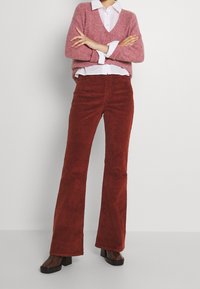 Pantalon évasé en velours côtelé rouge avec une texture douce, associé à un pull rose en maille porté sur une chemise blanche à col. Chaussures à talons carrés.