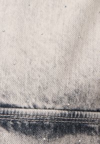 Gros plan d'un tissu en denim délavé avec des zones de lavage foncé et de petites clous argentés dispersés sur la surface près de la couture cousue.