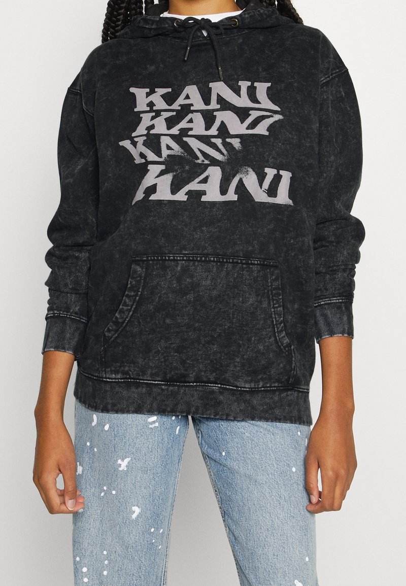 Personne portant un sweat à capuche noir délavé avec le texte répété "KANI" en blanc et un jean bleu clair avec des éclaboussures de peinture blanche, debout les mains le long du corps.
