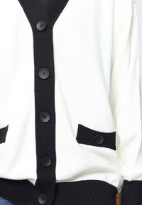 Cardigan blanc avec des accents noirs, présentant un col en V, six boutons noirs et deux poches latérales avec des poignets côtelés. Texture de tissu douce.