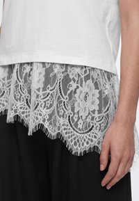Bordure en dentelle blanche avec motifs floraux sur l’ourlet d’un haut blanc porté sur un pantalon noir, avec une main droite visible à côté du corps.