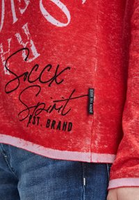 Rotes Langarmshirt mit weißem Druck, schwarzer "Soccx Spirit Est. Brand"-Text, einem Kleidungsetikett "Down the River", getragen mit blauen Jeans.