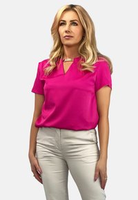 Fuksjowa bluzka z krótkimi rękawami ma dekolt w kształcie dziurki na klucz oraz gładkie satynowe wykończenie, zestawiona z jasnobeżowymi dopasowanymi spodniami.