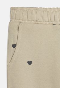 Beige bombažne hlače z ribastim elastičnim pasom, ki vsebujejo majhne črne srčne vzorce in stranske žepe. Gladka tekstura z casual dizajnom.