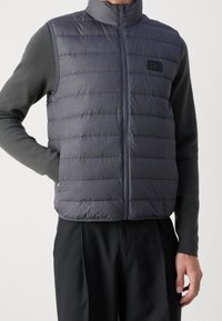 Szara pikowana kamizelka męska z wysokim kołnierzem, zamkiem błyskawicznym z przodu oraz naszywką z logo. Materiał wydaje się gładki; cechuje się lekką, ocieplaną konstrukcją.