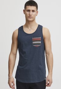 Tricou fără mâneci de culoare bleu, fabricat dintr-un material ușor, având un buzunar pe piept cu un tipar în zigzag de culori roșu, verde și albastru. Textură netedă.