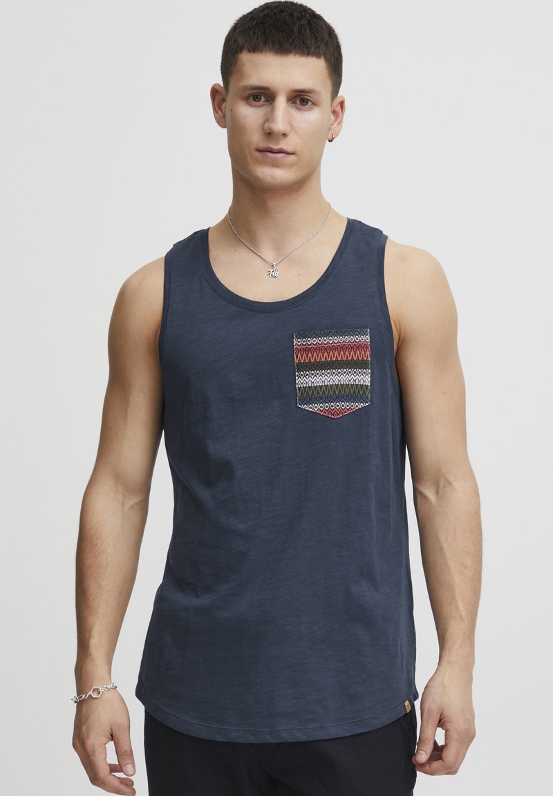 Tricou fără mâneci de culoare bleu, fabricat dintr-un material ușor, având un buzunar pe piept cu un tipar în zigzag de culori roșu, verde și albastru. Textură netedă.