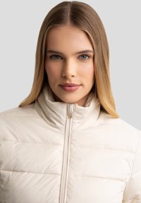 Letvægts, cremet pufferjakke med høj krave, frontlynlås, vandret quiltmønster og glat, blank finish.