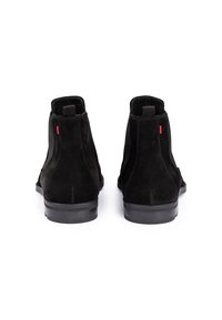 Par de botas de tobillo de ante negro con lengüetas y detalles de costura roja, mostradas desde la parte posterior sobre un fondo blanco.