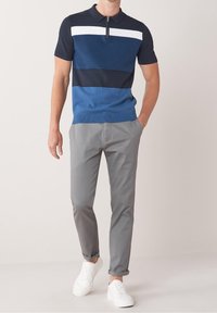 Navy and blue color-blocked polo shirt with zip collar, paired with light gray trousers and white sneakers. Smooth fabric, fitted design.
