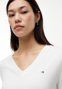 Jeune femme aux cheveux raides et foncés, portant des boucles d'oreilles anneaux argentés et un pull blanc à col en V avec un petit logo rouge, blanc et bleu sur la poitrine.