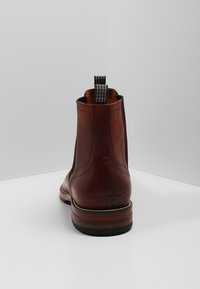 Botte Chelsea en cuir marron avec languette de tirage noire et blanche, design élégant, petit talon et semelle à rayures rouges. Finition texturée avec des détails subtils.