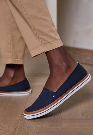 Donkere persoon die beige broek en marineblauwe instapschoenen met witte zolen draagt, staand op een houten vloer binnen.