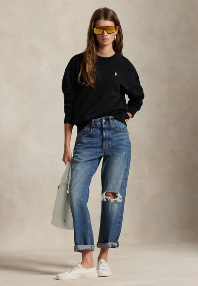 Kvinne iført svart genser, revne blå jeans, hvite joggesko, store gule solbriller, holder en hvit veske, stående mot en ensfarget bakgrunn.