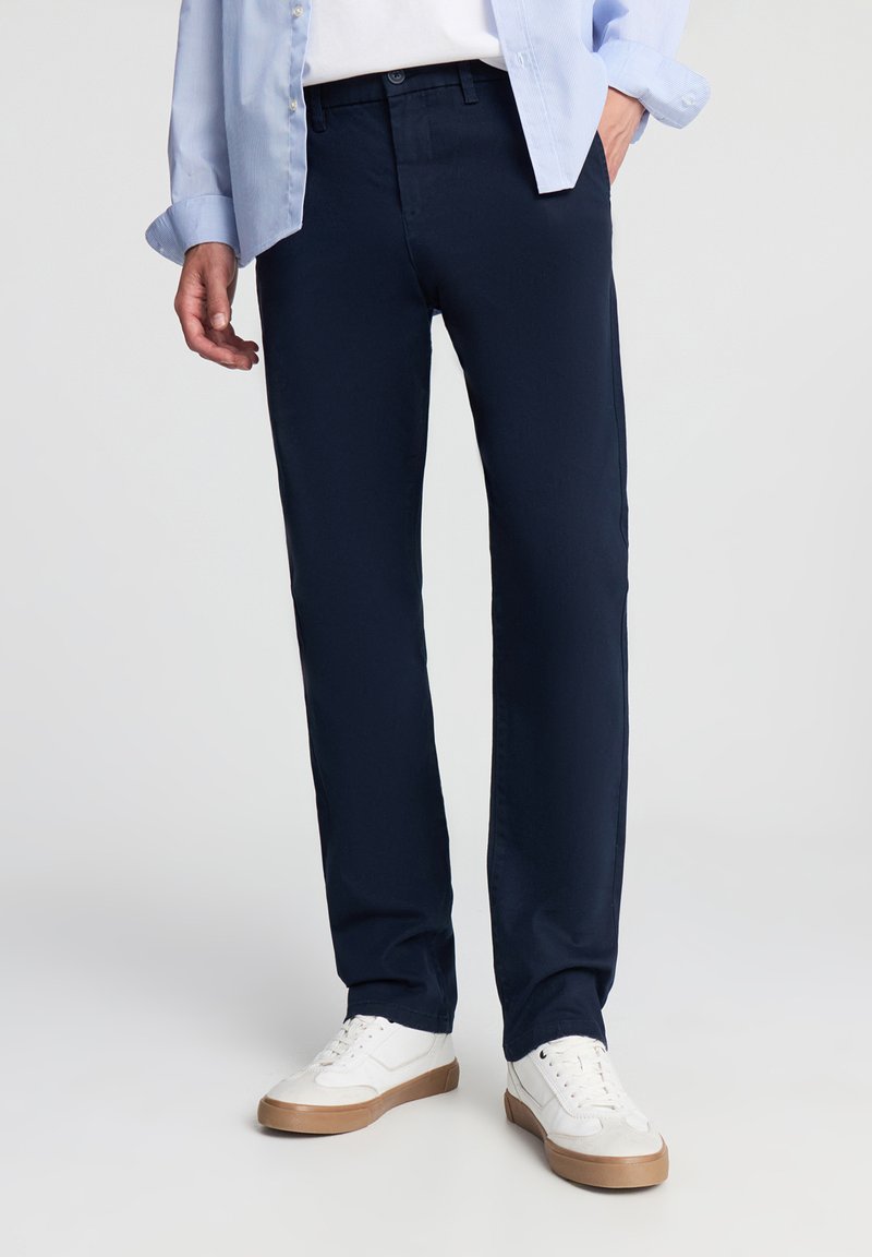 Pantaloni blu navy con una texture liscia, design a gamba dritta e passanti per cintura. Abbinati a una camicia azzurra e sneakers bianche.