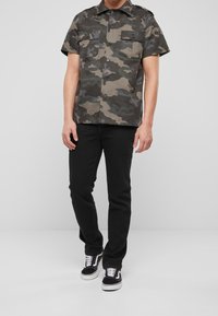 Camicia a maniche corte in camouflage con due tasche sul petto, chiusura con bottoni, realizzata in tessuto leggero; abbinata a jeans neri e sneaker nere.
