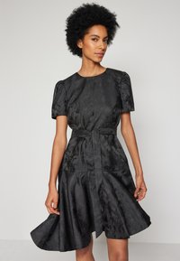 Μαύρο σατέν φόρεμα με κοντά φουσκωτά μανίκια, εφαρμοστό κορσέ και φουσκωτή φούστα μέχρι το γόνατο. Διαθέτει ζώνη στη μέση και υφασμάτινο σχέδιο.