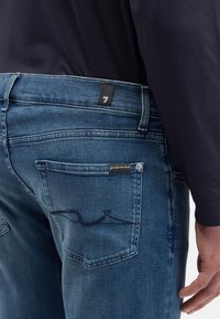 Męskie niebieskie jeansy z denimu z tylną kieszenią ozdobioną haftowanym wzorem, czarną metką z numerem 7 oraz subtelną przetartą fakturą.