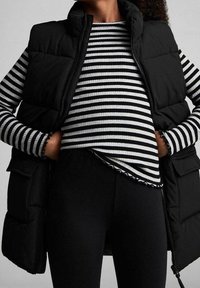 Czarna kamizelka puchowa na czarno-białej pasiastej bluzce z długim rękawem, o prążkowanej fakturze i dopasowanym kroju. Czarne legginsy na dole.