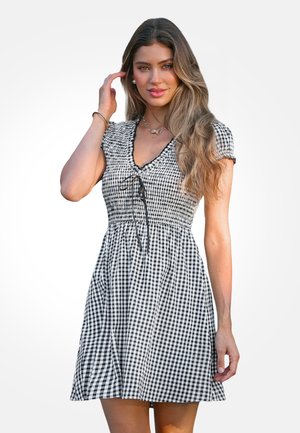 Młoda kobieta z długimi włosami, ubrana w krótką sukienkę w czarno-białą kratę, dotykająca włosów i patrząca przed siebie.