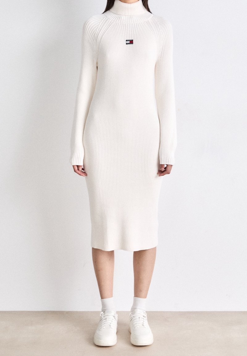 Femme portant une robe midi en maille côtelée blanche avec col roulé et manches longues, associée à des chaussettes blanches et des baskets blanches.