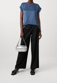 Haut en satin bleu avec manches retroussées, pantalon noir à jambes larges, sac à main métallique argenté avec bandoulière chaîne, porté avec des mules noires à bout ouvert.