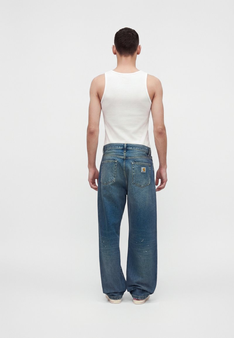 Homme portant un débardeur blanc et un jean bleu ample, debout de dos, devant un fond blanc uni.