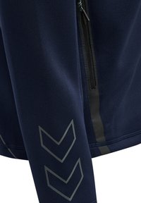 Jakkeærme i marineblå med to grå chevron-striber; tilstødende sort lynlåslomme på jakkens krop synlig.