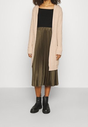Femme portant un gilet à capuche beige, un haut noir, une jupe midi plissée vert olive et des bottines noires épaisses sur un fond uni.