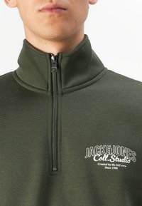 Grøn fleecepullover med høj krave, frontlynlås og hvidt logotekst. Glat tekstur og afslappet design.