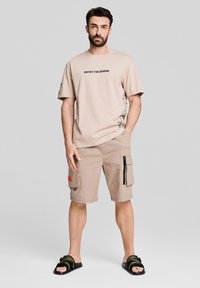 Beżowy t-shirt z czarnym napisem "WEJDŹ DO LEGENDY" i grafiką. Beżowe spodenki cargo z kieszeniami na suwak. Czarno-zielone klapki.