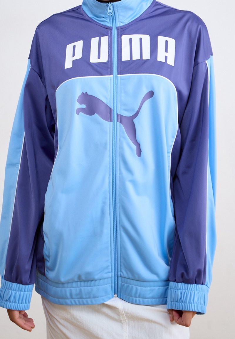 Ljusblå och lila Puma sportjacka med dragkedja, Puma-logotyp och text framtill, bärs av en person med avslappnade händer vid sidorna.