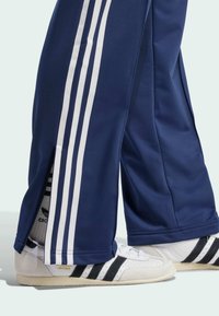 Navy track pants with white stripes and side zip, paired with white sneakers featuring black stripes and gold "Japan" detail.